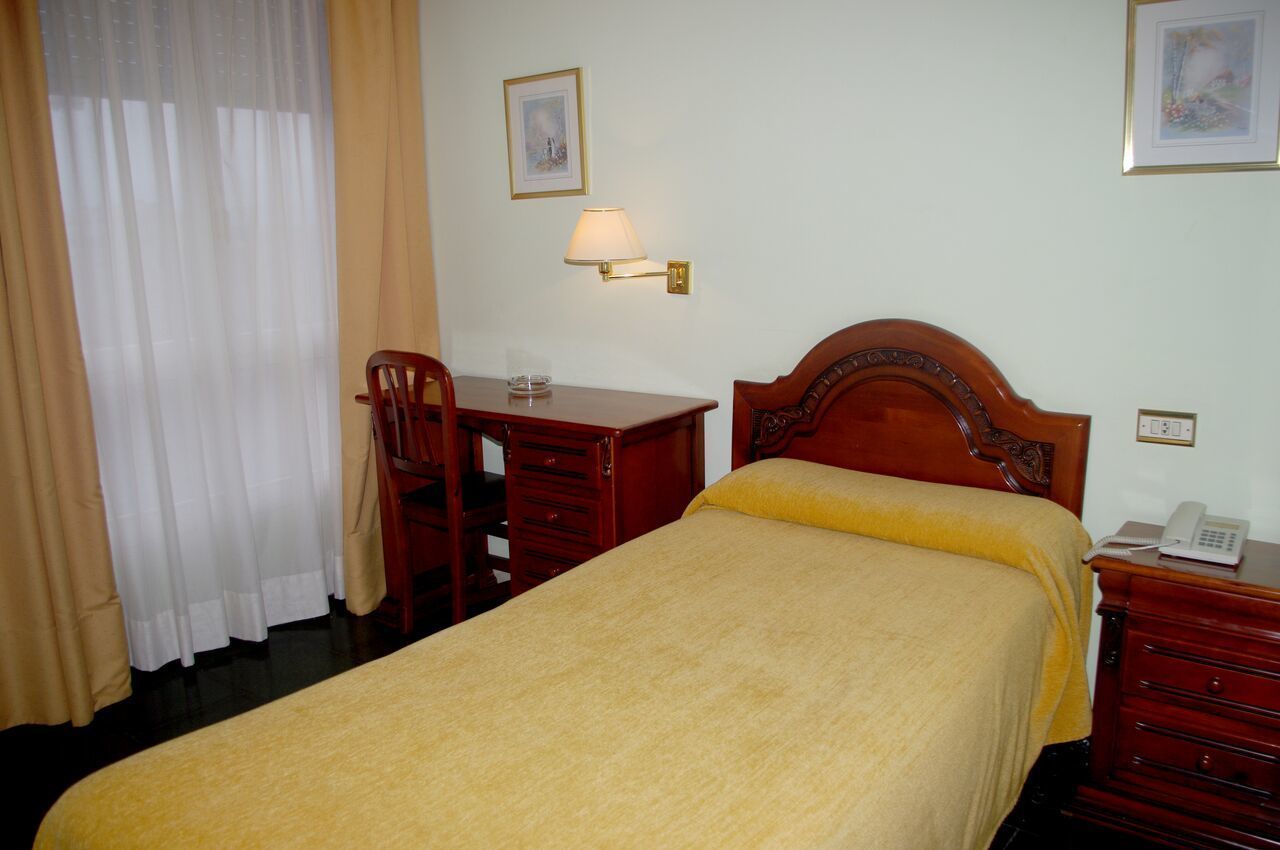 A hotel room with a bed, a desk with a lamp, a chair, and a telephone on a nightstand.