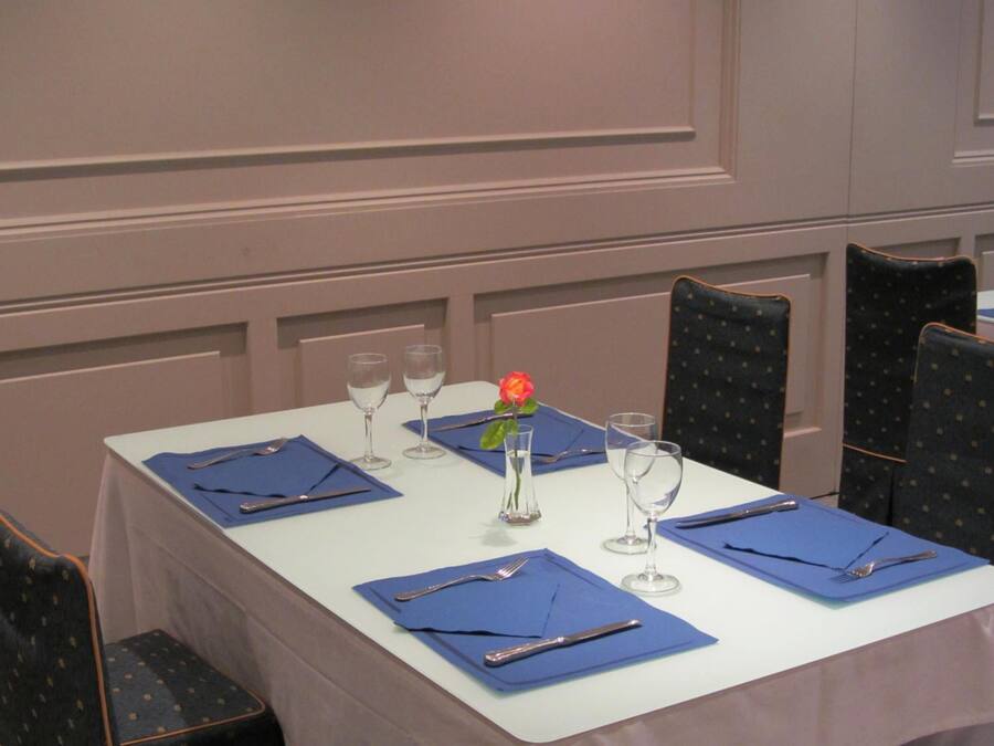 Una mesa de comedor puesta con servilletas azules, cubiertos y copas de vino, con un florero que contiene una sola flor.