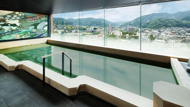 Une piscine intérieure moderne avec des parois en verre transparent, une terrasse en bois et une vue sur le paysage urbain et les montagnes.