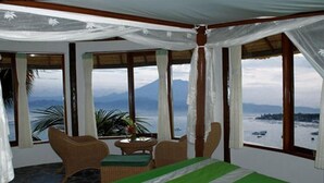 A room with a bed, a table, and two chairs. There are large windows with a view of the sea and mountains. - Coconut Beach Resort (BALI)