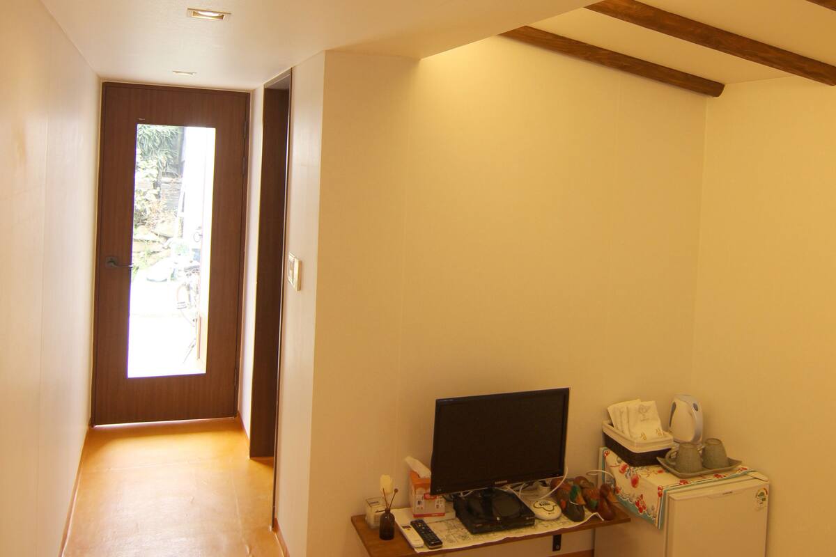 Un couloir étroit, avec une porte en bois au fond, un petit bureau avec un ordinateur et un réfrigérateur, et un luminaire au plafond.