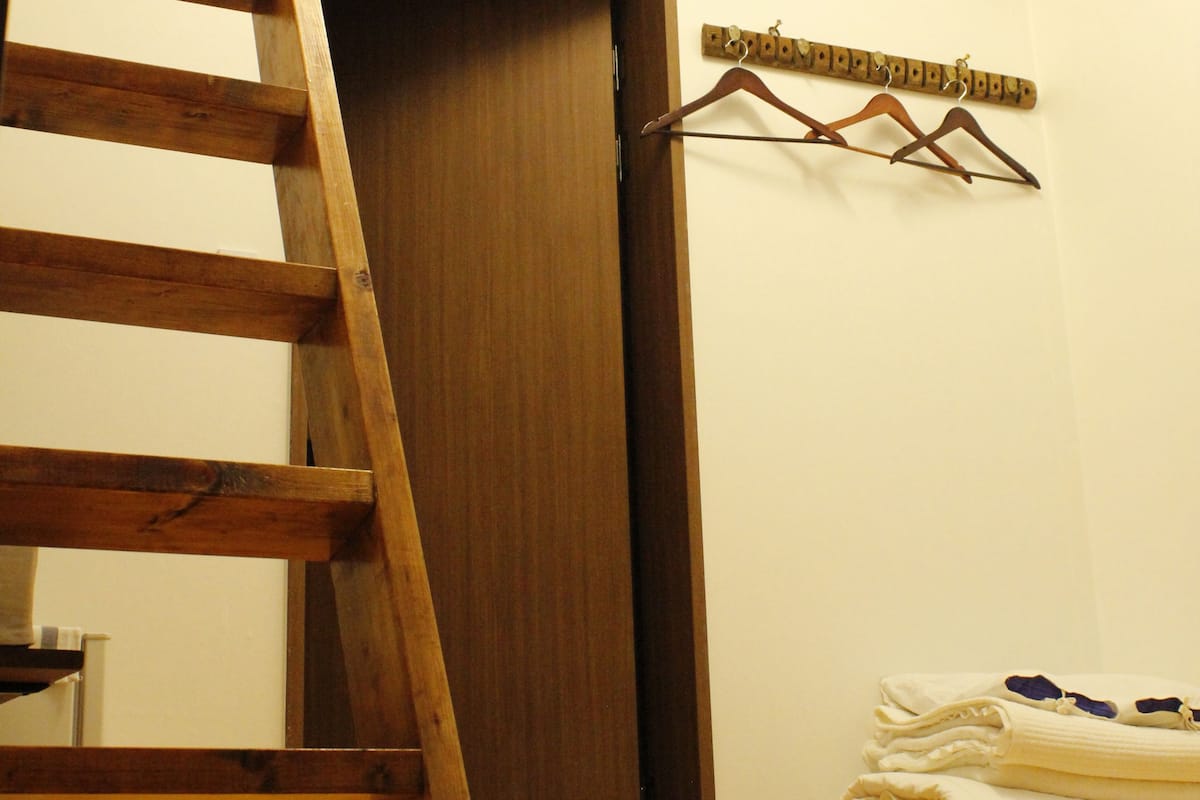 Un escalier en bois mène à une chambre située en mezzanine, avec une rambarde en bois, une porte et un porte-manteau fixé au mur.