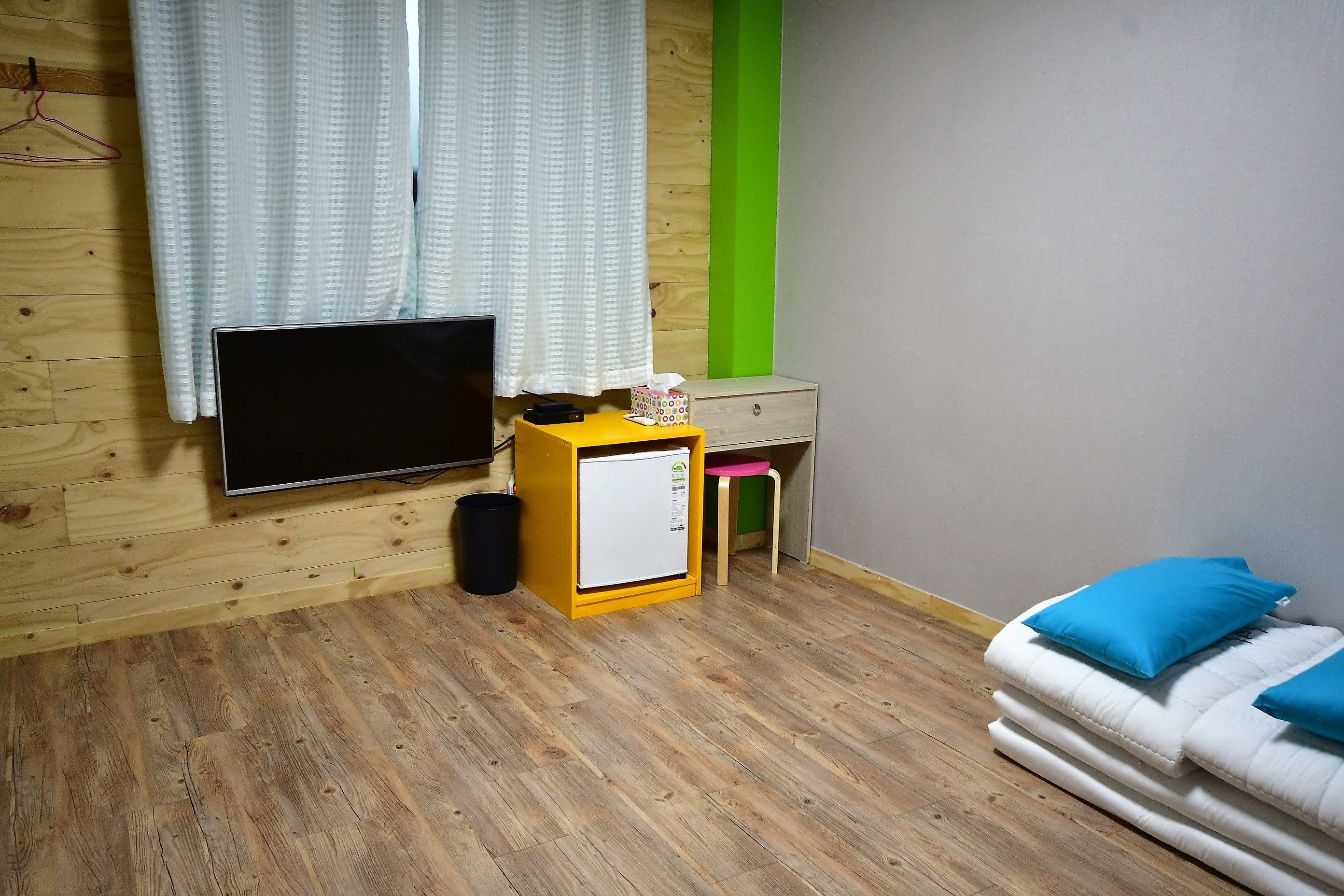Une pièce avec un plancher en bois, un téléviseur fixé au mur, une petite armoire jaune, un bureau avec une chaise et un lit avec des oreillers bleus.