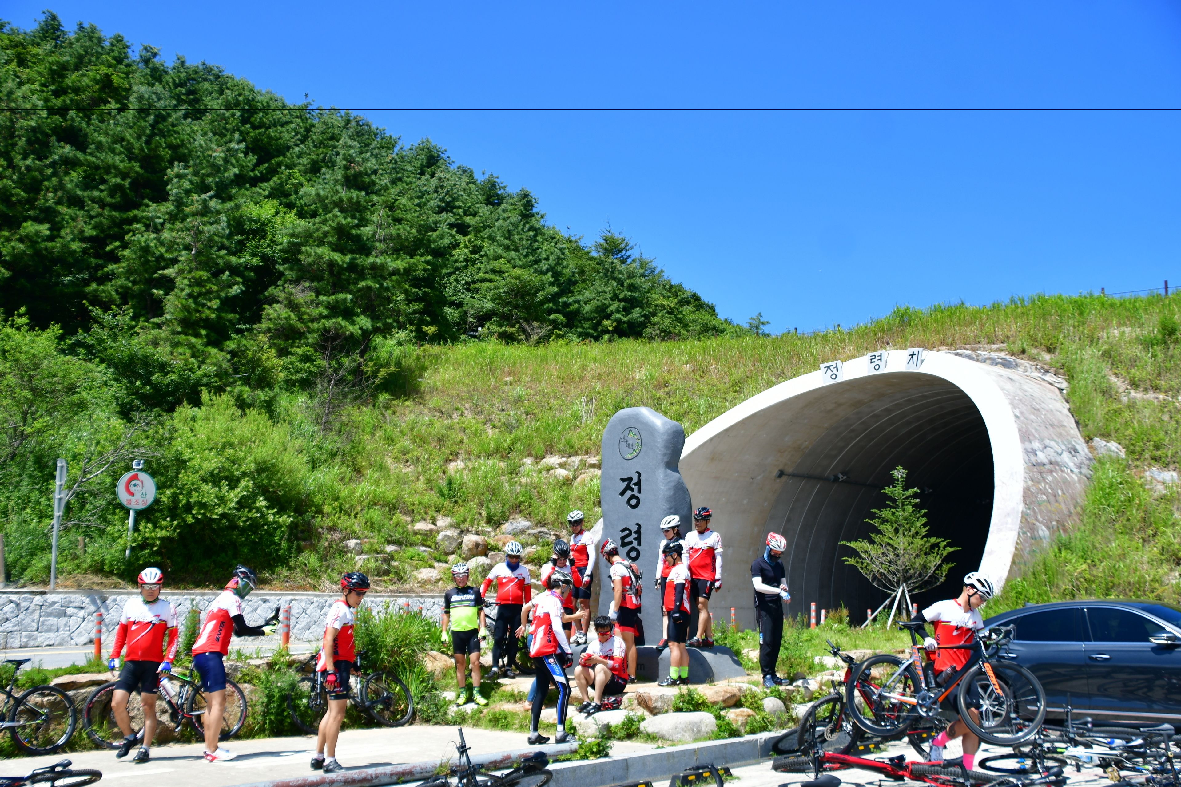 A group of cyclists at a tunnel entrance with a stone monument, surrounded by greenery and a clear sky.
