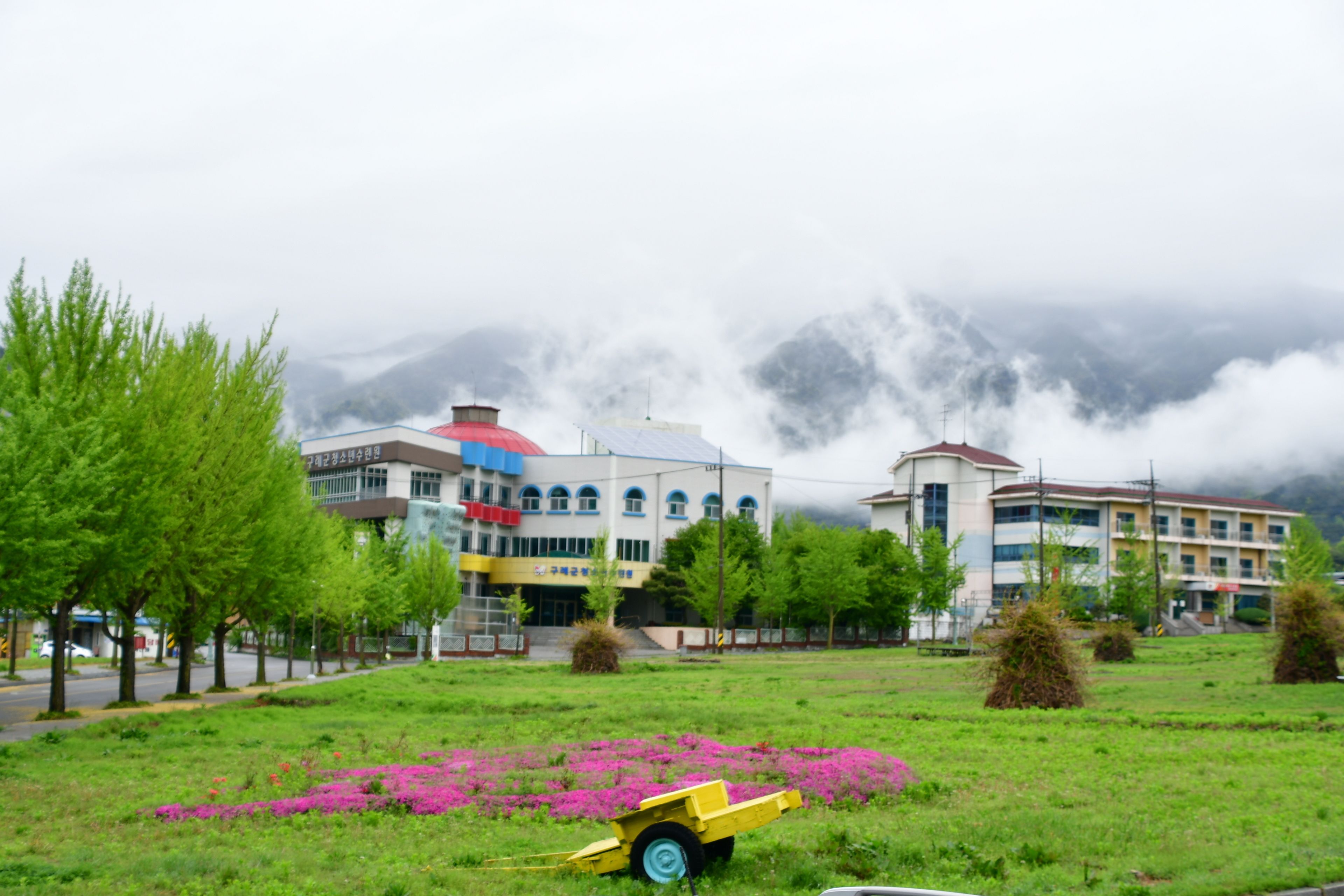 A grassy area with a yellow cart, buildings with multiple stories, and a mountainous backdrop.