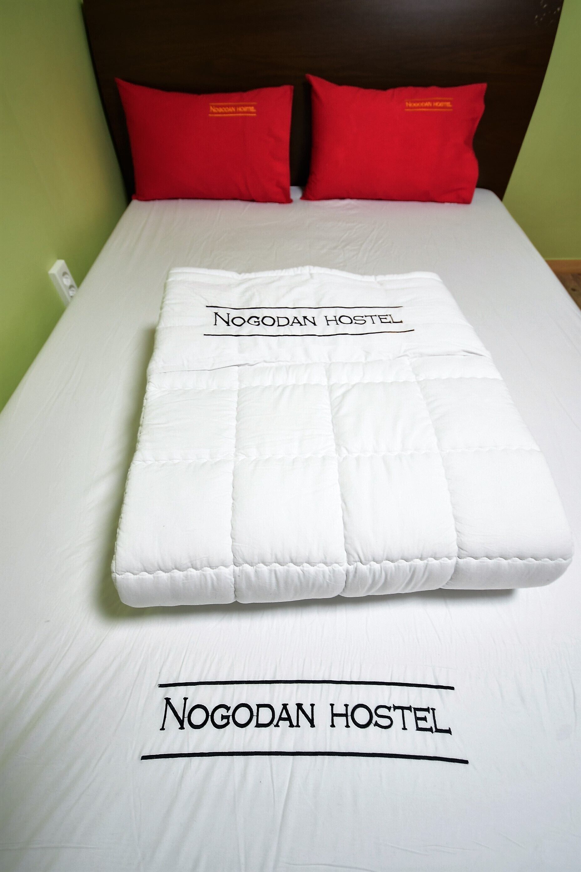 Un lit bien fait, recouvert d’une couette blanche et agrémenté de deux oreillers rouges, avec une étiquette indiquant « Auberge de Nogodan ».