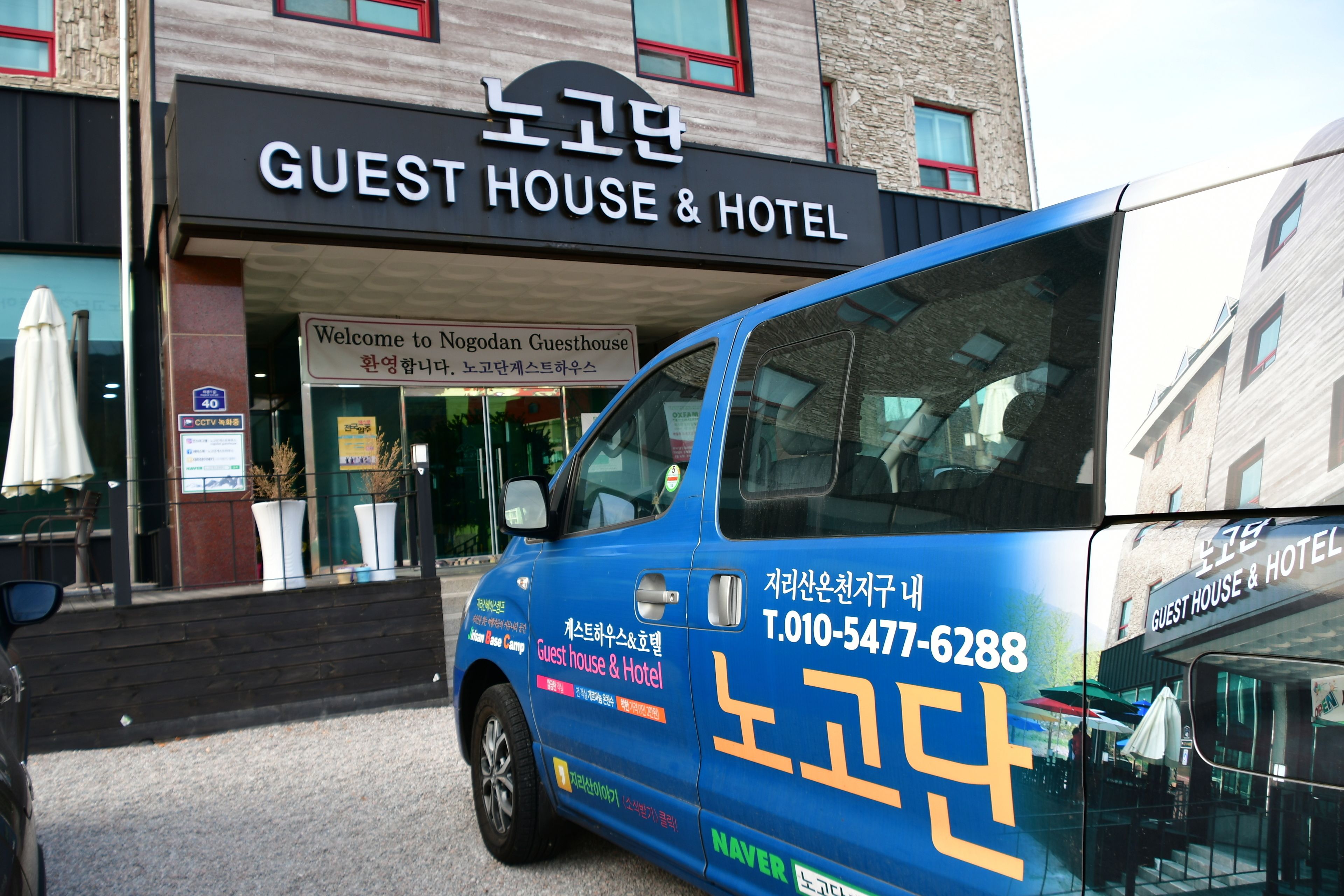 Una camioneta azul estacionada frente a un edificio con el letrero "GUEST HOUSE & HOTEL", con un cartel que da la bienvenida a visitantes a Nogodan Guesthouse.
