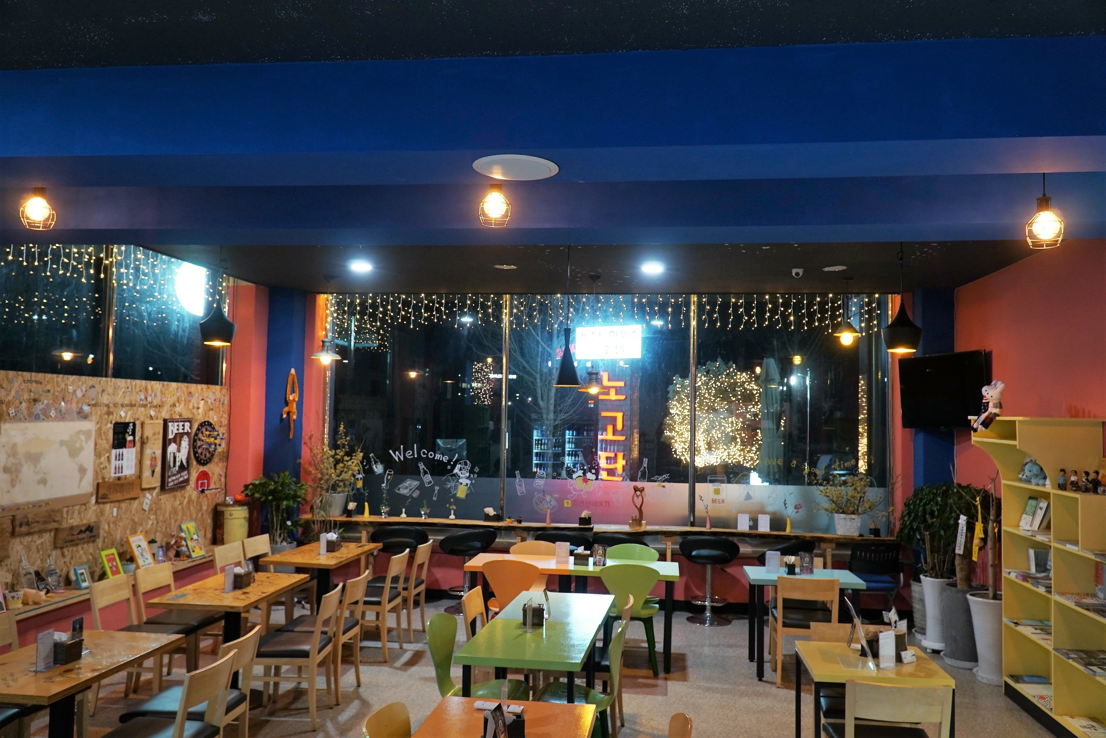 Un accogliente ristorante con tavoli e sedie di legno, un'area bar e un'ampia finestra decorata con luci a stringa.