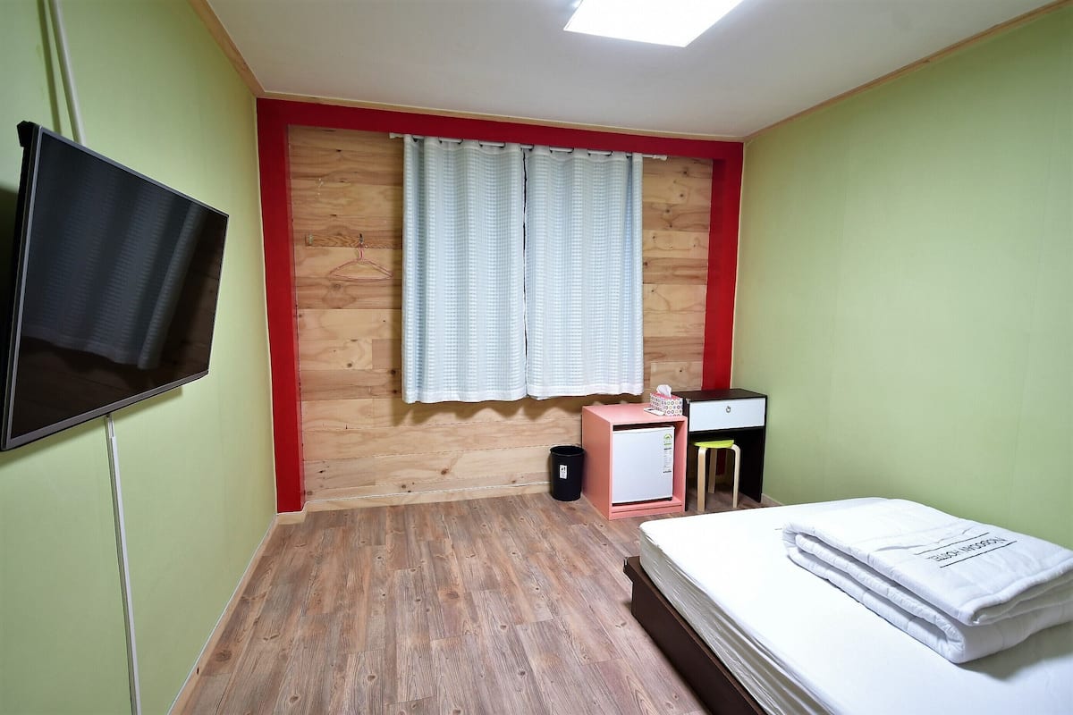 Ein kleiner, ordentlicher Raum mit einem Bett, einem Fernseher, einem kleinen Schreibtisch und einem Fenster mit Vorhängen.