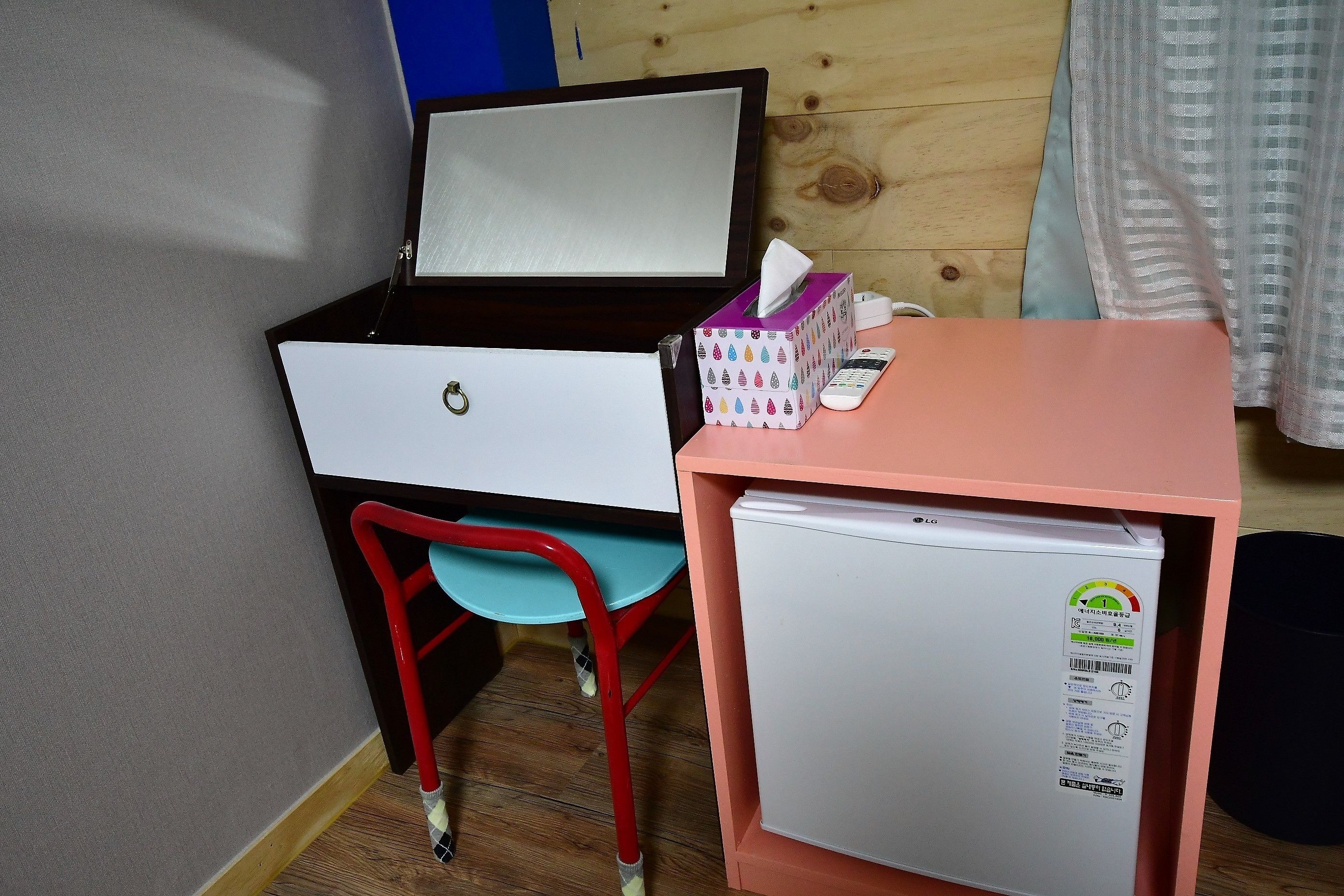 Een kleine kamer met een bureau, een stoel, een kleine koelkast en een zakdoekendoos.