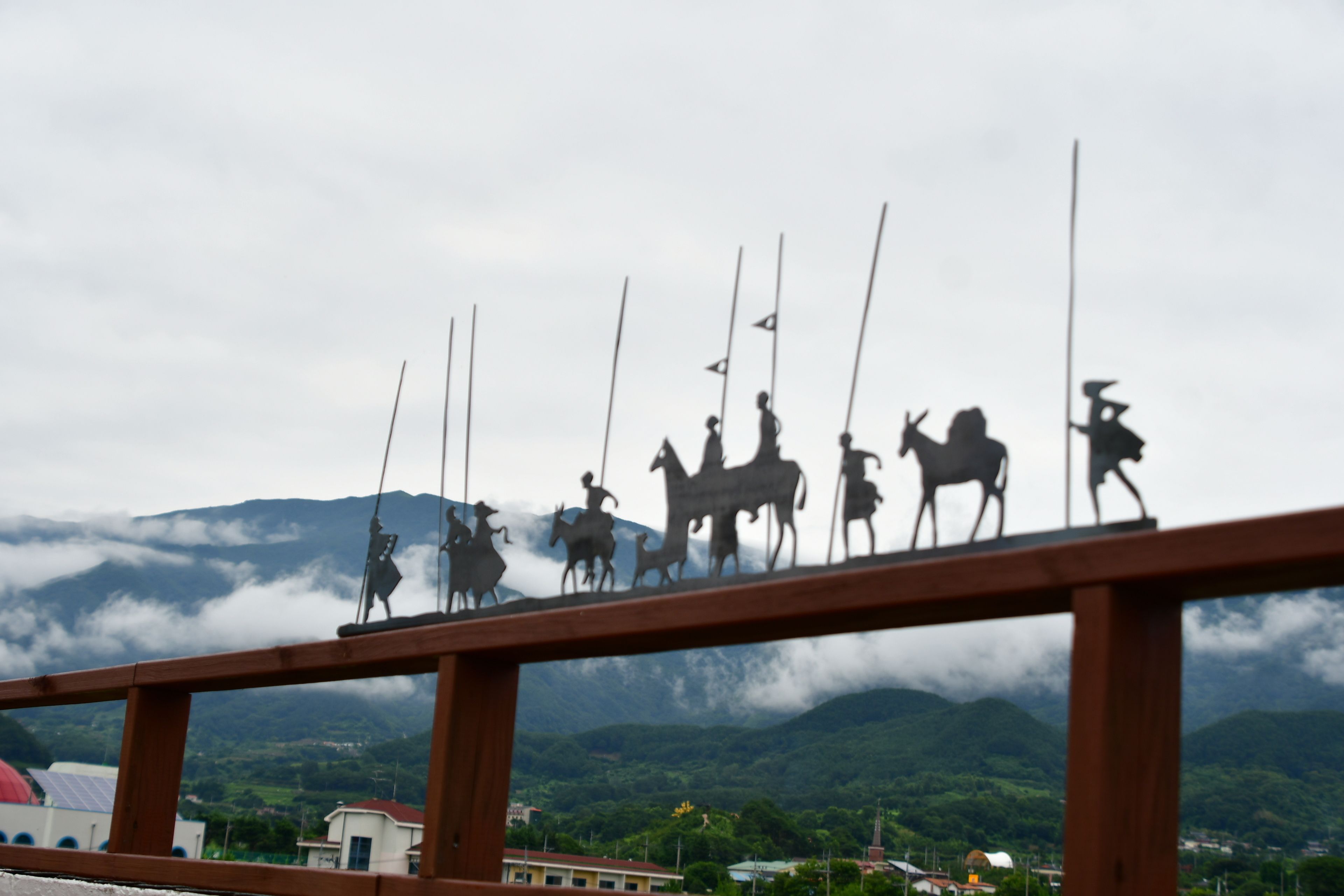 Blick auf eine Stadt mit einer markanten Skulptur von Personen zu Pferd, einer Kirche und Bergen im Hintergrund.