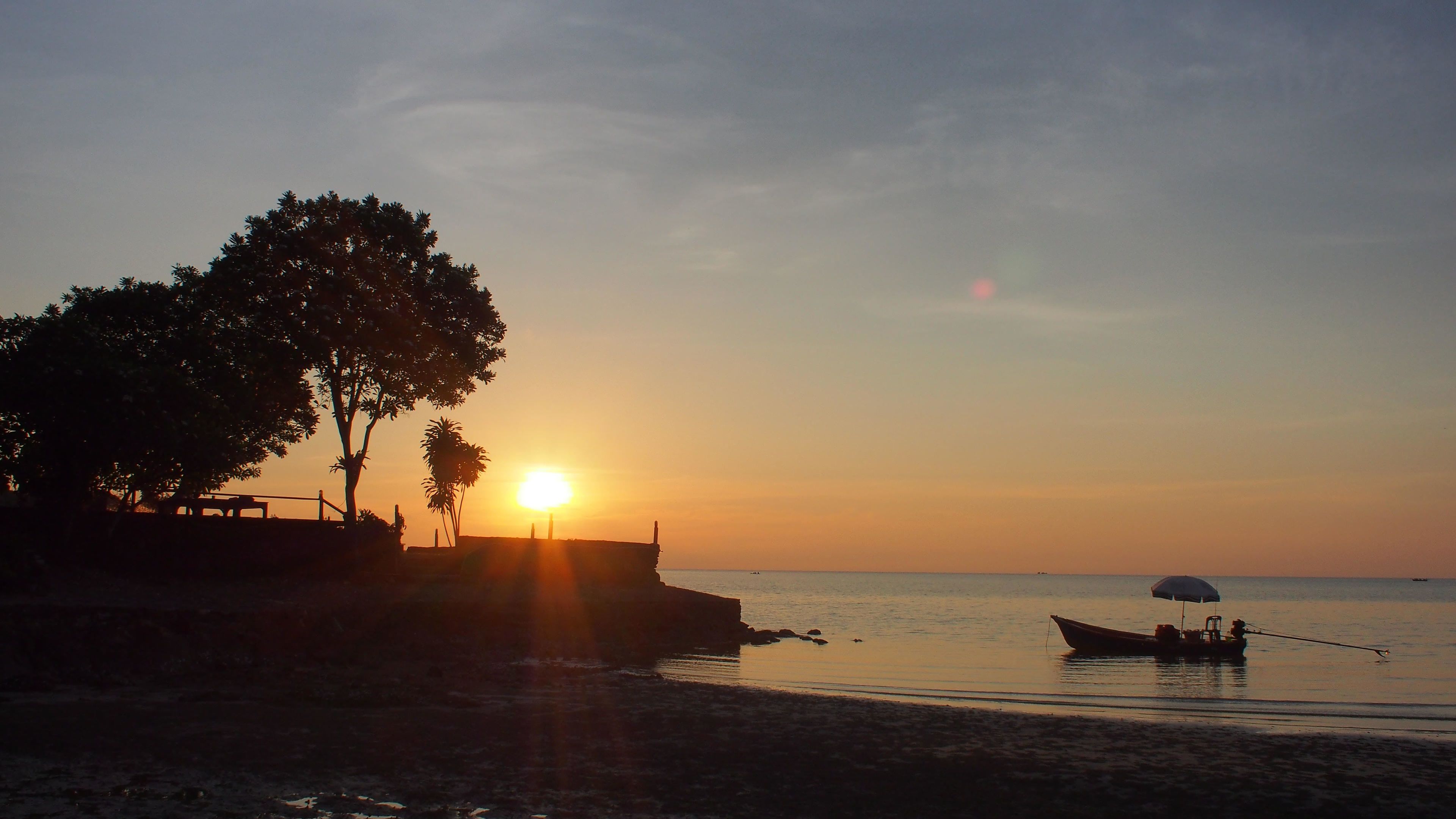 Un atardecer sobre una playa con la silueta de un bote y un árbol.