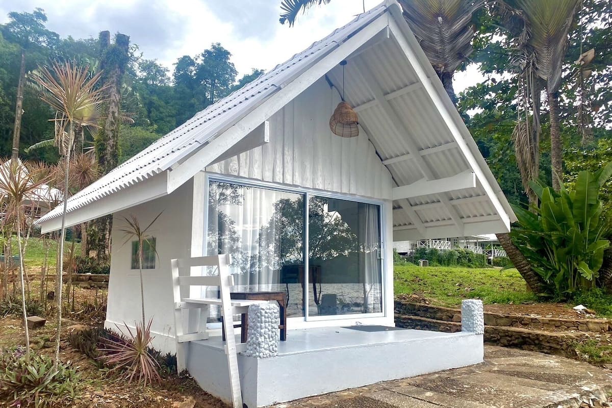 Uma casa pequena e branca, térrea, com telhado inclinado, grandes janelas de vidro e varanda de concreto.
