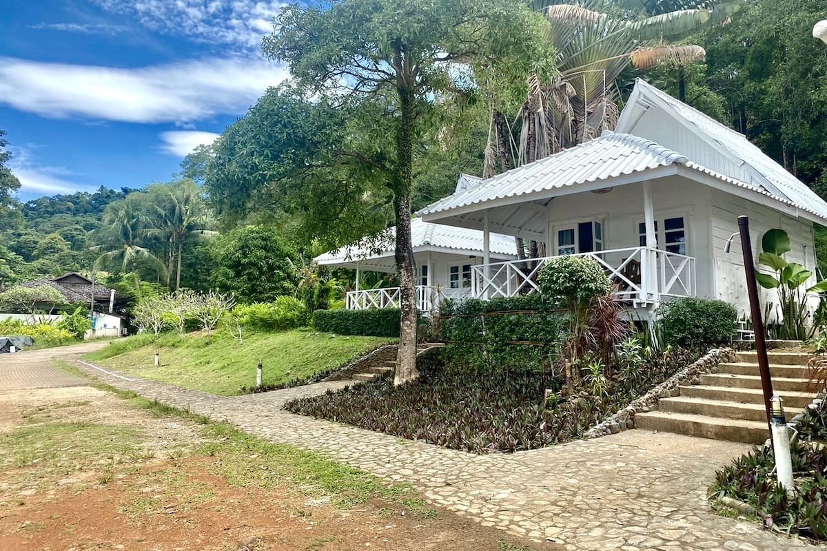 Um edifício branco com varanda coberta, rodeado por vegetação exuberante e um caminho de pedra que leva à entrada.