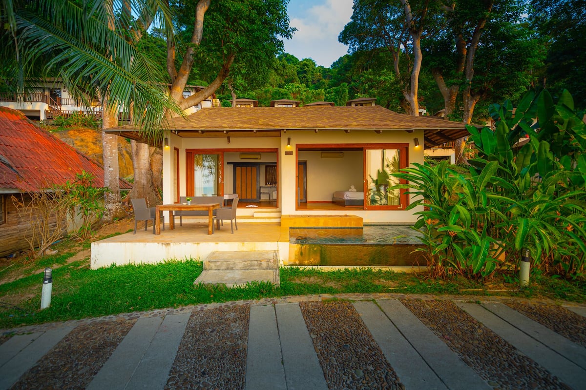 Uma casa de dois andares com um pátio, uma piscina pequena e muita vegetação.