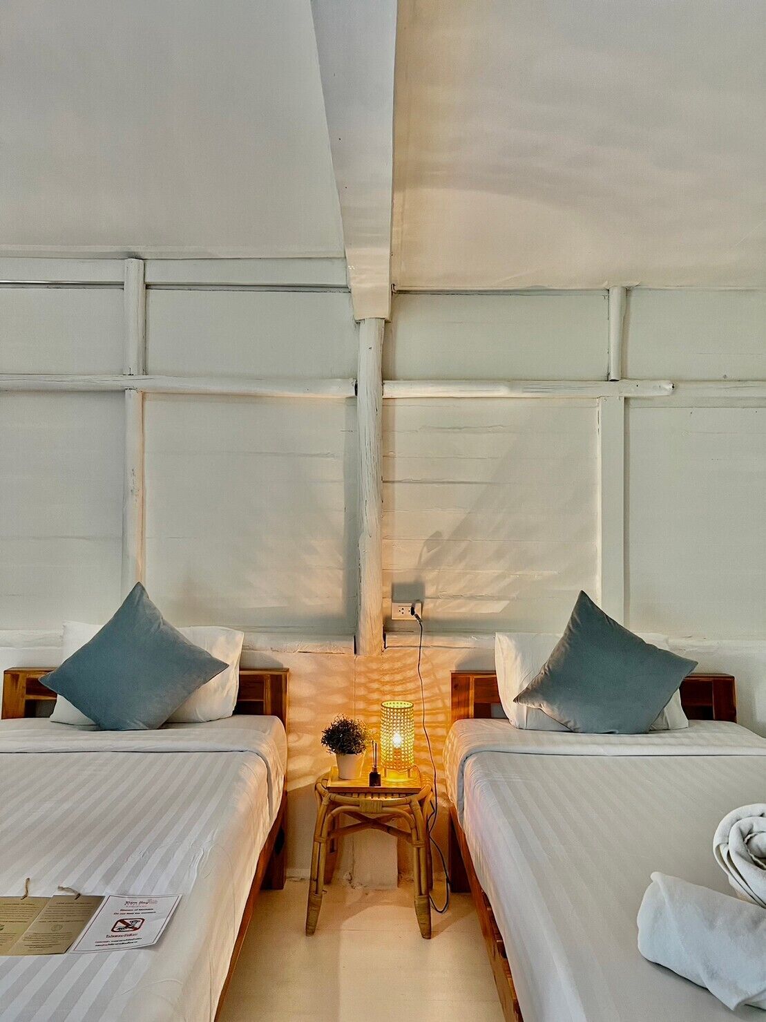 Habitación de hotel con dos camas, cada una con una almohada y una mesita de noche. En el centro de la habitación hay una mesita con una lámpara.