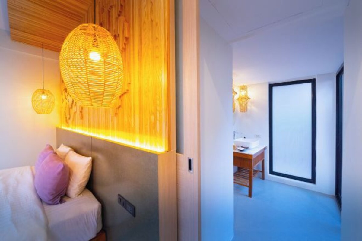 A modern hotel room with a bed, a wooden headboard, a large woven light fixture, and a view of the bathroom.