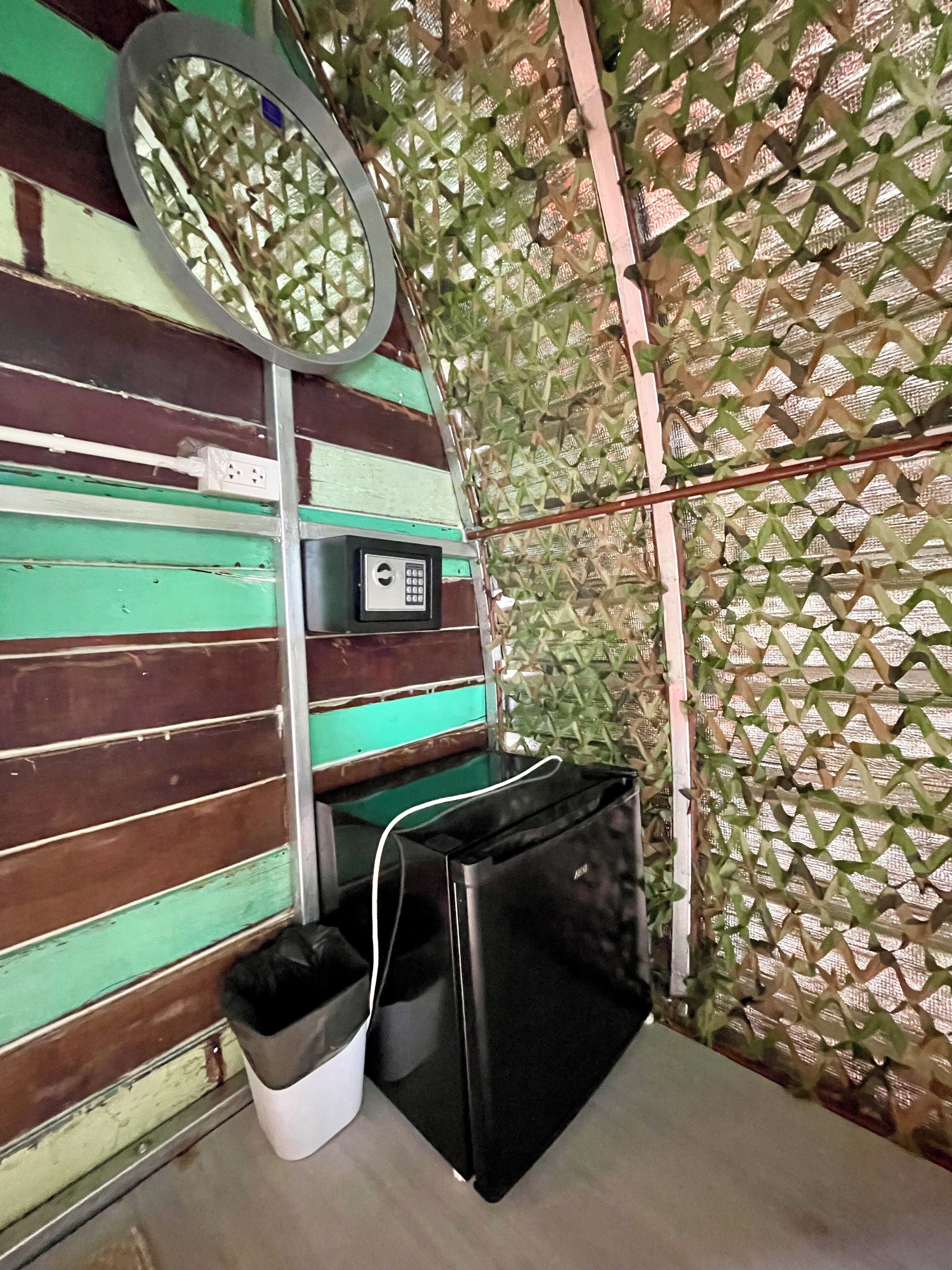 A room with a green and brown striped wall, a small window with a circular mirror, a wall-mounted electrical outlet, and a black appliance on the floor.