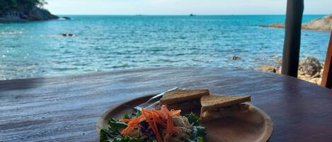 A wooden table with a plate of food and a sandwich, overlooking a body of water with a distant shoreline.