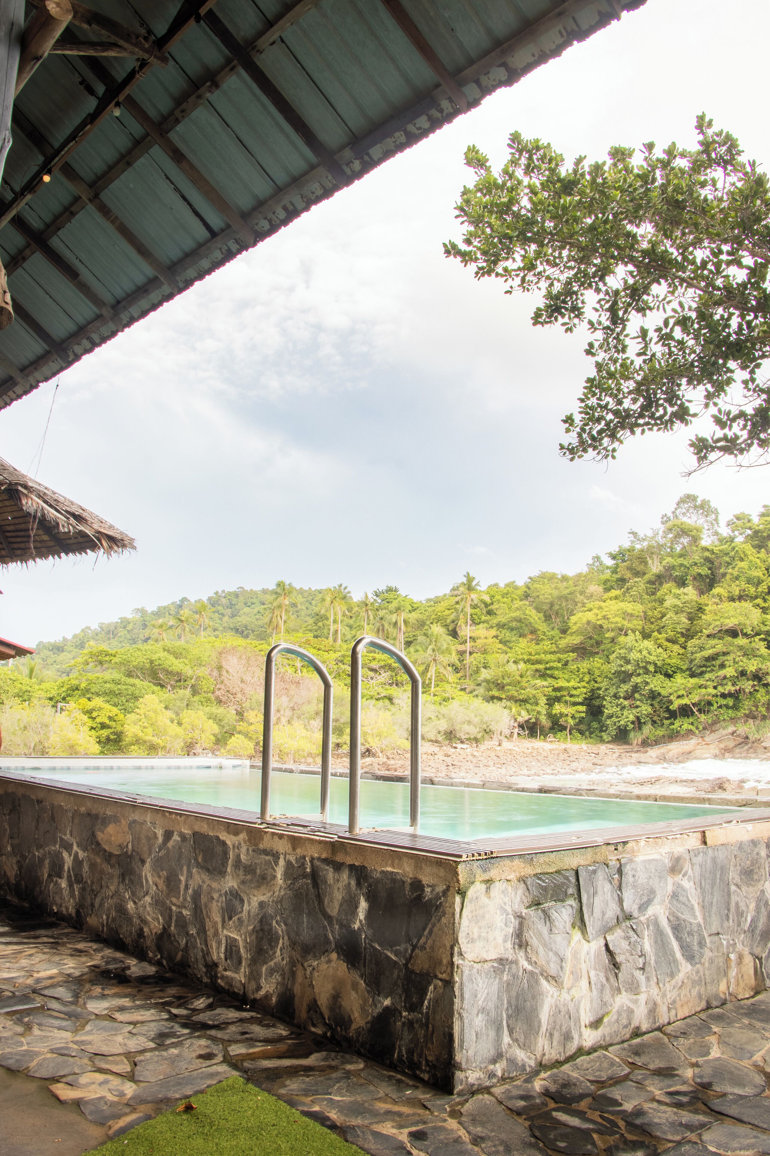 Una piscina de piedra con escaleras metálicas, rodeada por un muro de piedra y una estructura de techo de paja.
