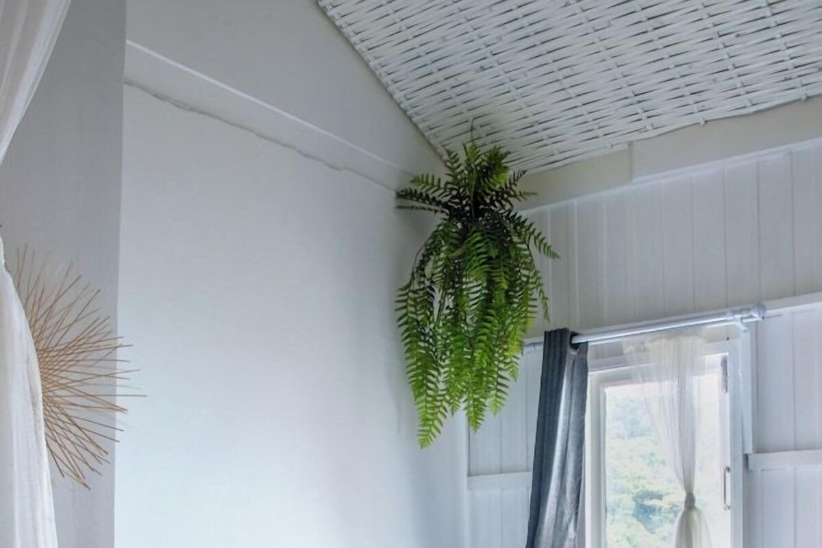 Cama bem arrumada com lençóis brancos, planta pendurada, cadeira com almofada e janela com cortinas.