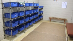 A storage room with metal shelves holding blue baskets, a wooden bench, and a wall calendar. - Hot Spring Hotel Boston (Obihiro)