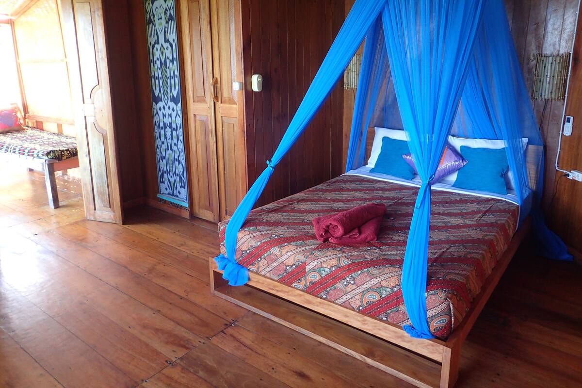 Een houten kamer met een bed onder een blauw hemelkleed, een houten bankje en een kamerkleed met dessin.