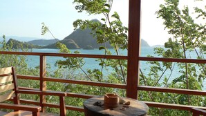A wooden deck with a bench and a table, overlooking a body of water and a mountain. - Waecicu Eden Beach Hotel (Labuan Bajo)