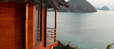 A wooden cabin with a thatched roof overlooking calm waters and distant islands.