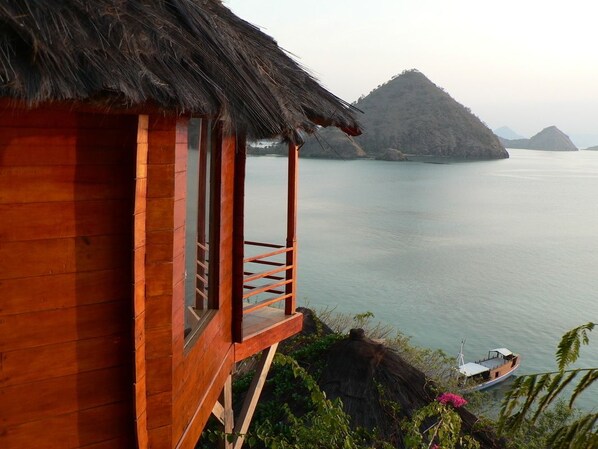 A wooden cabin with a thatched roof overlooking calm waters and distant islands.