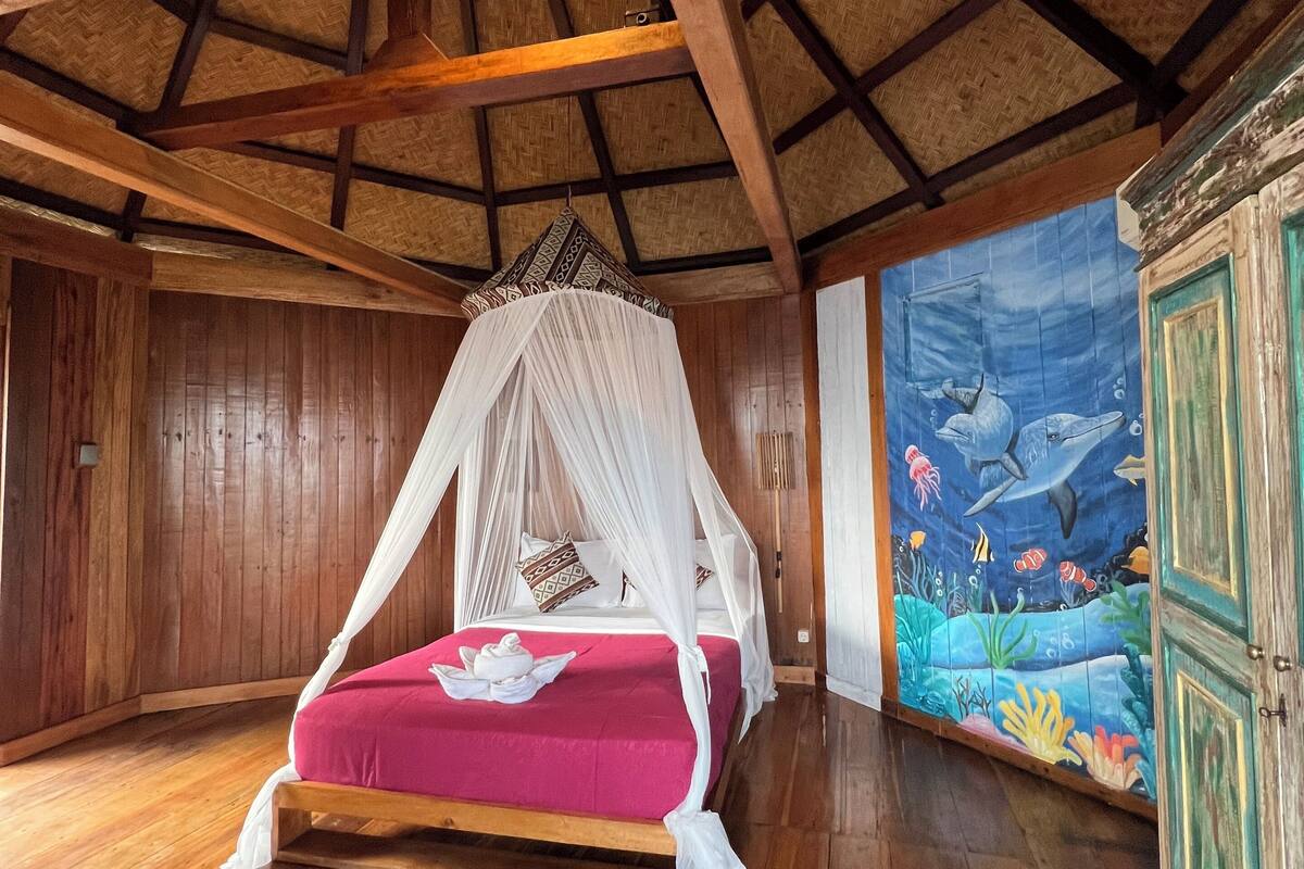 Een kamer met een houten vloer, een bed met hemelbed, een muurschildering van zeewezens en een houten kast.