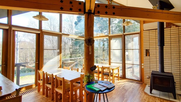 A spacious room with large windows, wooden furniture, and a wood-burning stove. - Pension Gooseberry (Furano)