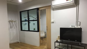 Una habitación con piso de madera, un televisor negro sobre un pedestal negro, un muro de vidrio con imágenes, y un aire acondicionado en la pared.