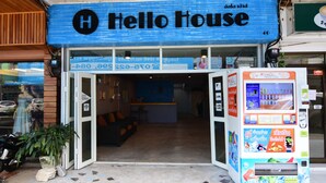 Entrada de hotel con un letrero azul que dice "Hello House", un mostrador de recepción y una máquina expendedora.
