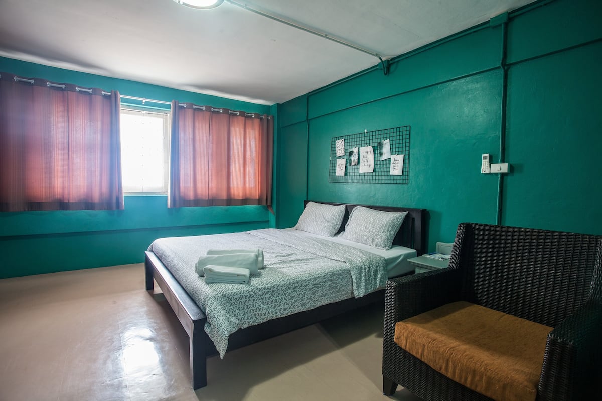 A bedroom with a bed, a chair, a window with curtains, a wall-mounted phone, and a bulletin board with notes.
