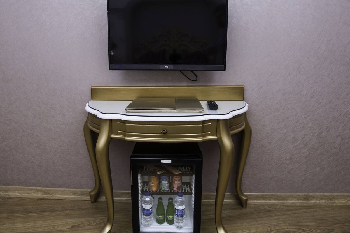 A room with a television mounted on the wall, a small table with a laptop and remote control, and a mini-fridge with beverages.