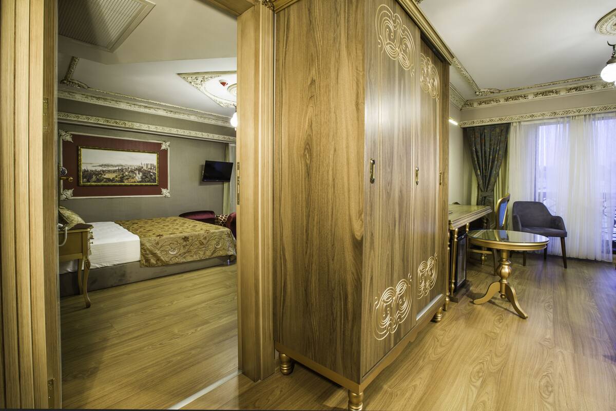 A hotel room with a wooden wardrobe, a bed with a patterned bedspread, a desk with a chair, and a framed picture on the wall.