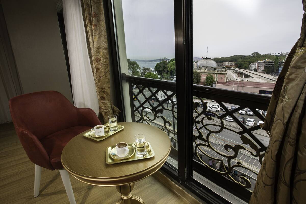 A room with a red chair, a small round table with a tray containing a cup and a glass, and a view of a cityscape through a balcony with ornate railings.
