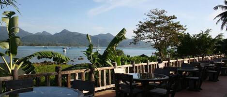 An outdoor dining area with tables and chairs, overlooking a body of water and mountains.