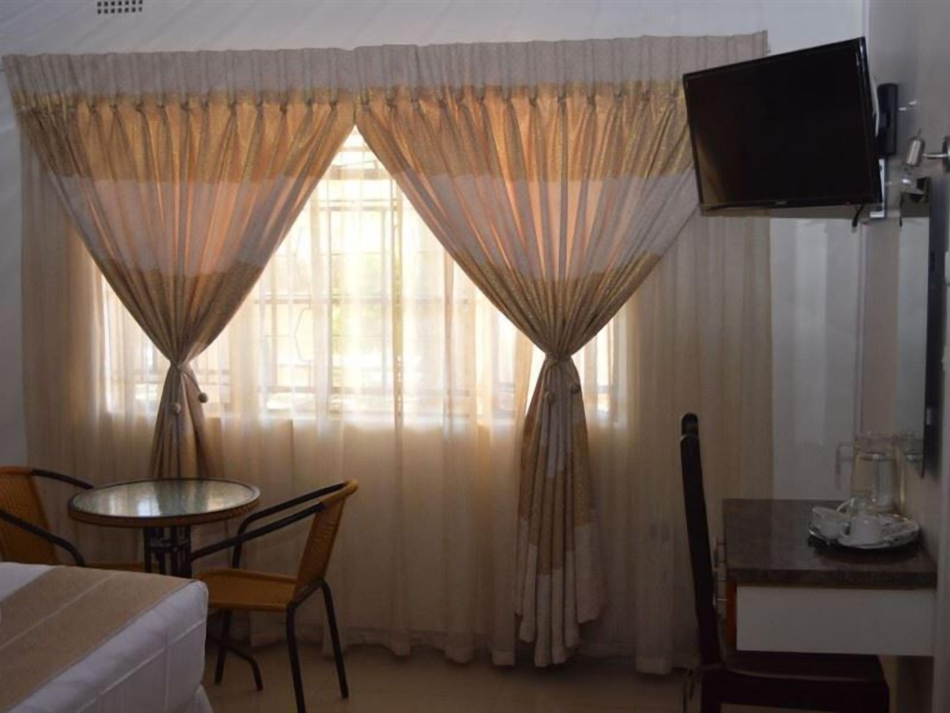 A hotel room with a bed, a small round table with chairs, a TV mounted on the wall, and a window with curtains.