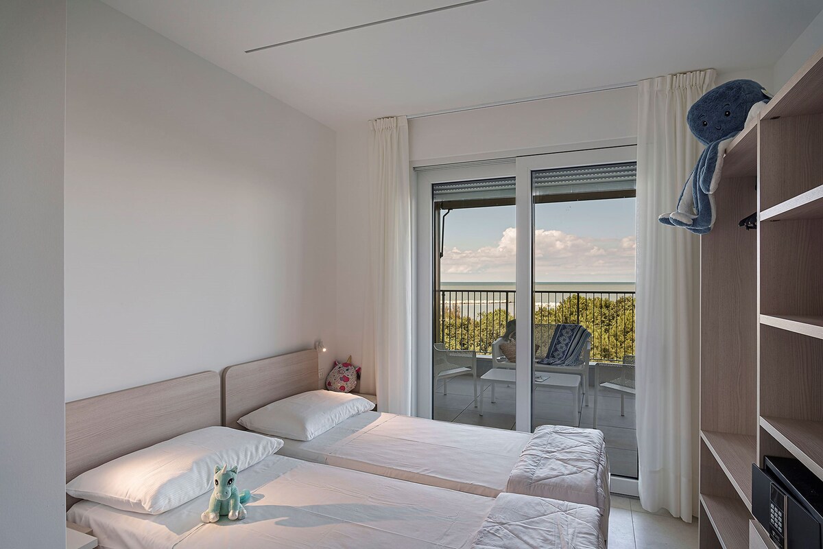 A hotel room with two beds, a balcony with a table and chairs, and a large window with curtains.