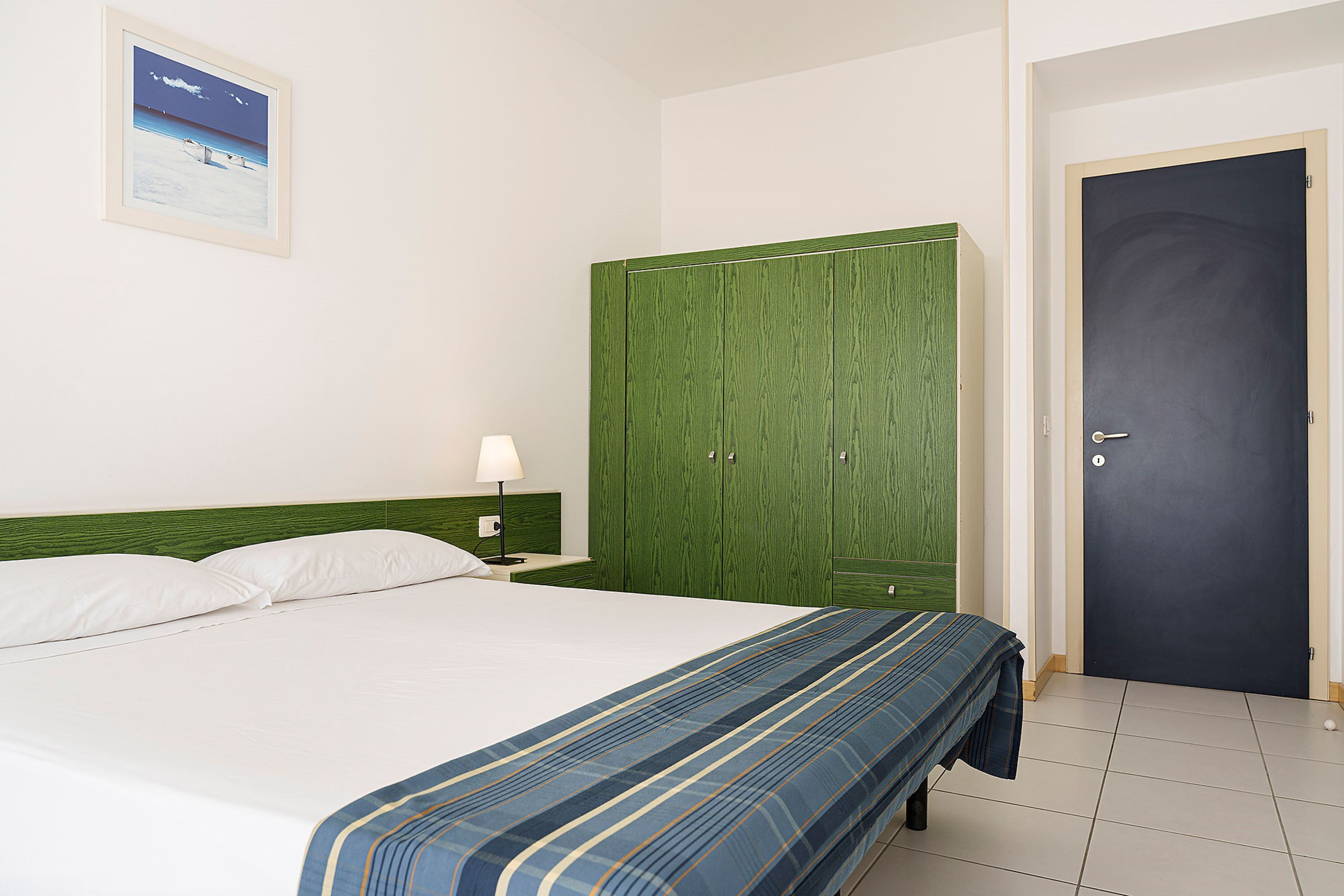 A hotel room with a bed, a green wardrobe, a door, and a framed picture on the wall.
