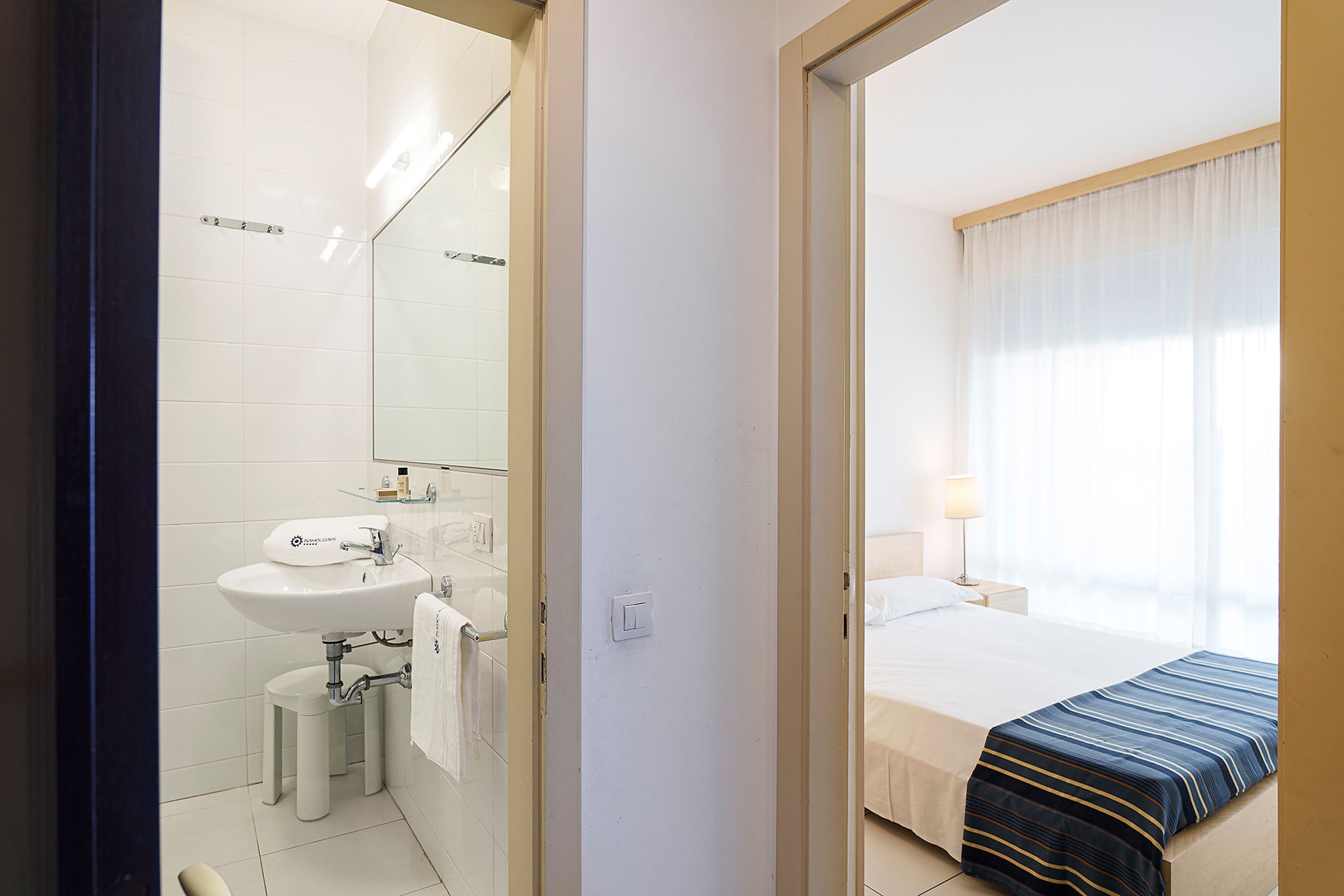 A hotel room with a bed, a bedside table, a lamp, and a bathroom with a sink and mirror.