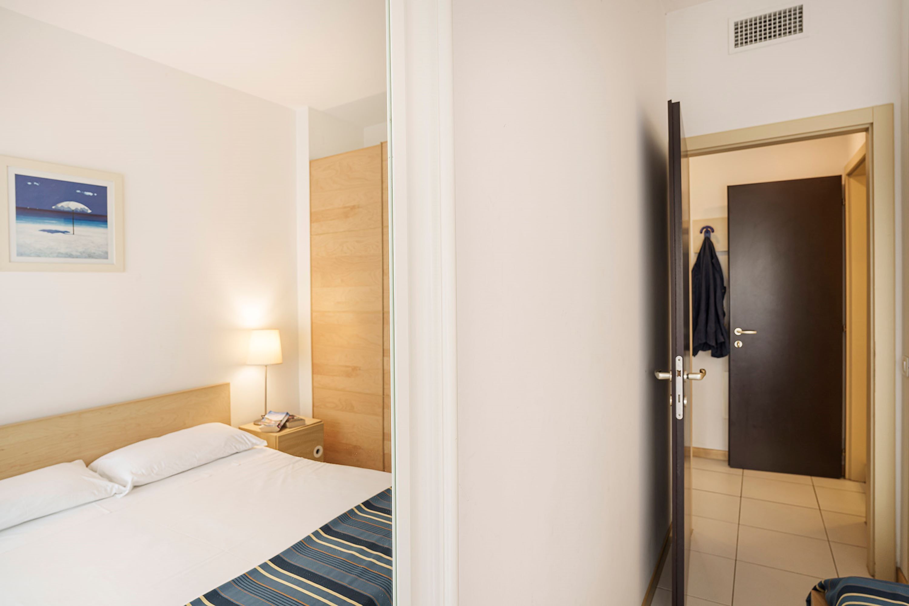 Habitación de hotel con cama, mesita de noche con lámpara, pared de paneles de madera y puerta que da a otra habitación.