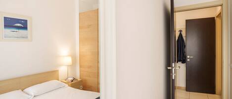 Habitación de hotel con cama, mesita de noche con lámpara, pared de paneles de madera y puerta que da a otra habitación.