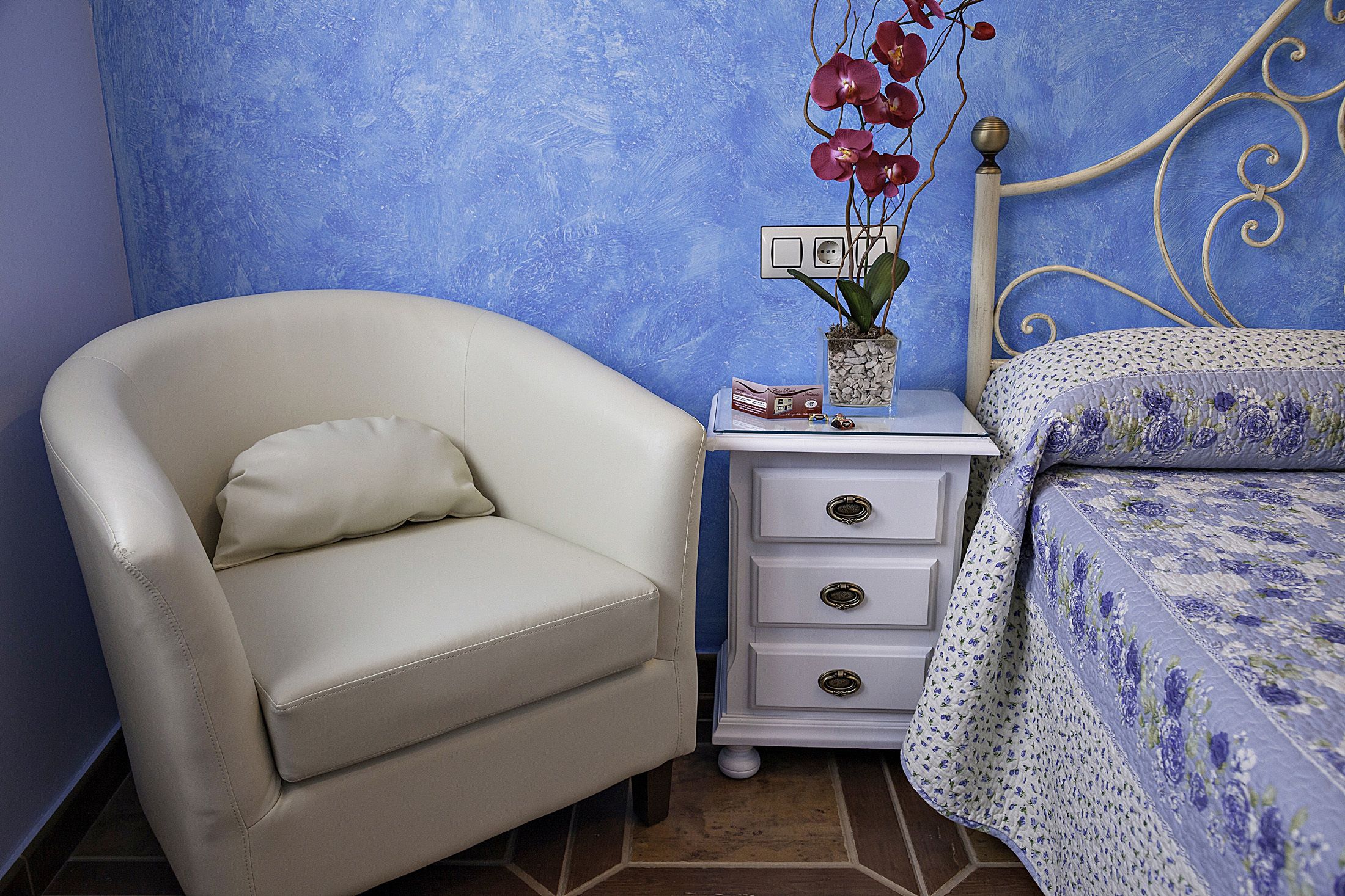 A bedroom with a white chair, a bedside table with drawers, a bed with a floral bedspread, and a blue textured wall.