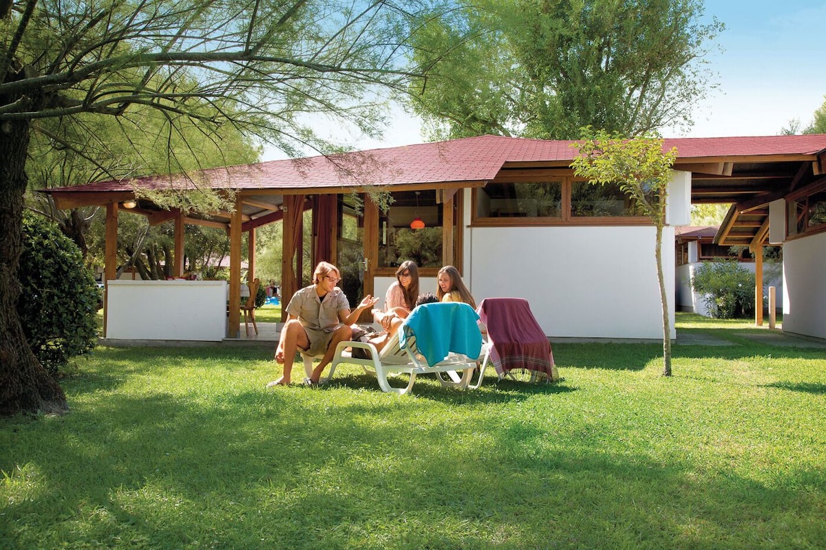 A covered outdoor seating area with a wooden structure and a red roof, surrounded by greenery and trees.