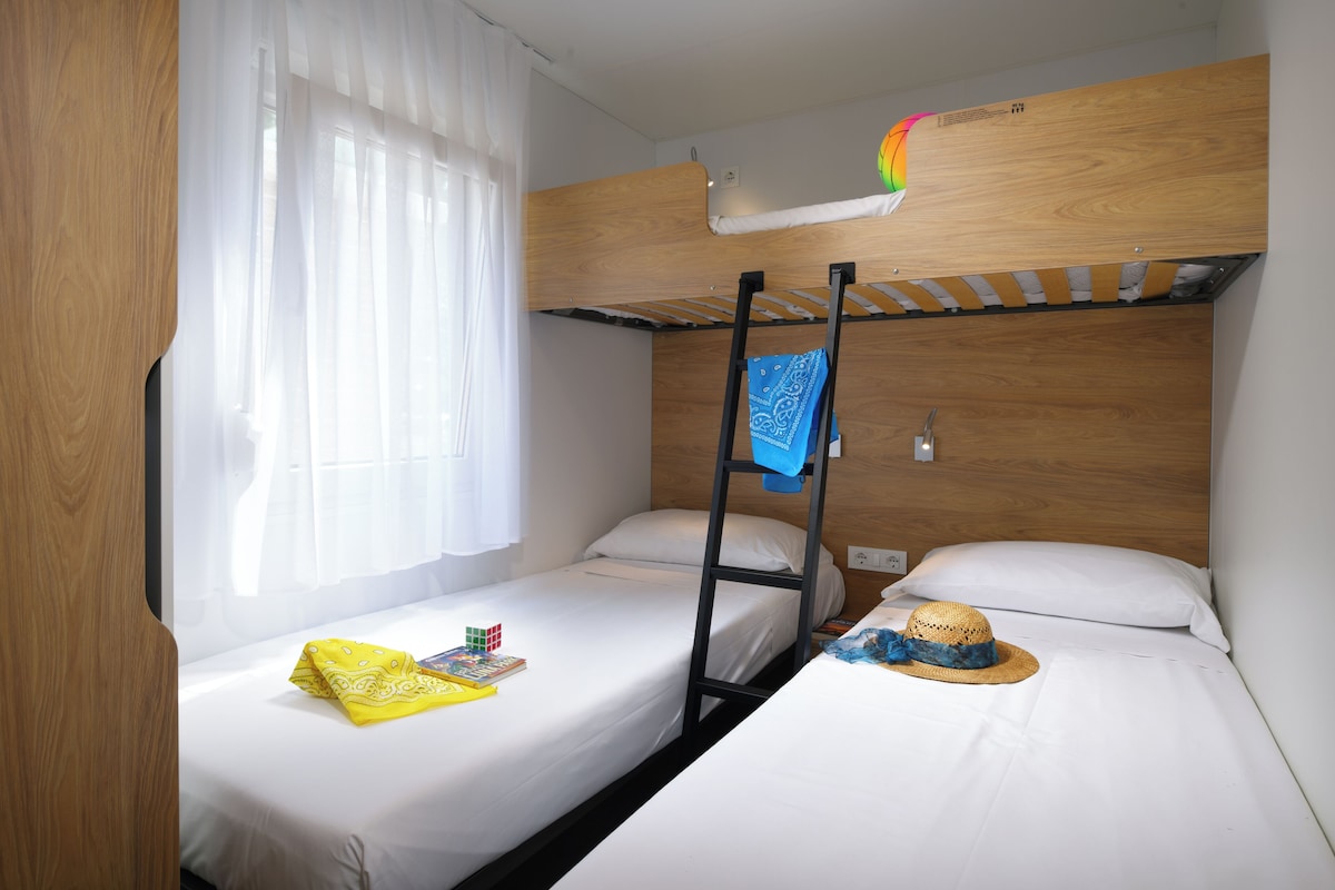 Une chambre avec un lit superposé, une échelle, une fenêtre avec des rideaux légers et un chapeau posé sur l’un des lits.