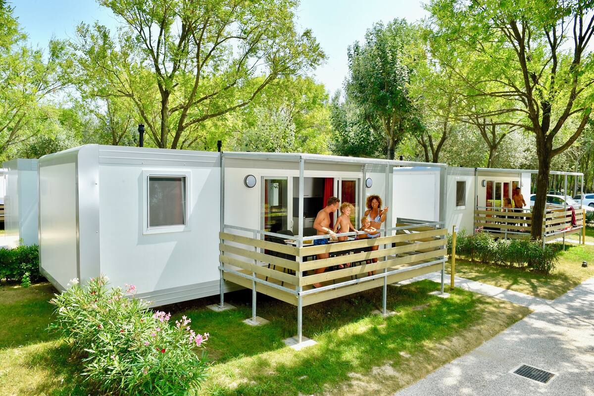 A white mobile home with a balcony, surrounded by greenery and trees.