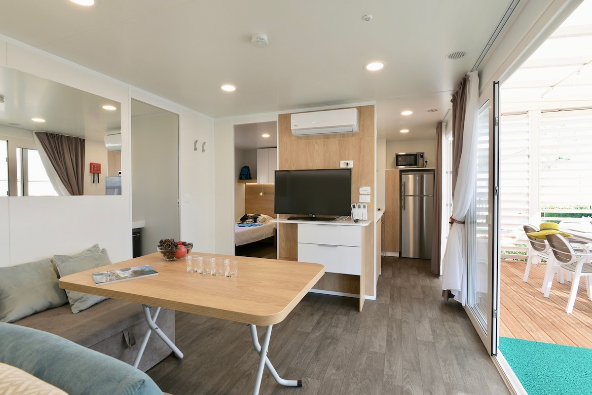 Un espace de vie moderne et compact, comprenant une table à manger en bois, un canapé, un téléviseur, un réfrigérateur et une porte-fenêtre coulissante donnant sur un patio.